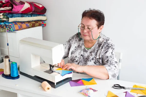 Costura y acolchado en el taller de una mujer sastre - mujer mayor con gafas trabaja en un escritorio con una máquina de coser e hilo, telas, agujas, alfileres, botones, tijeras, cortadores rotativos . — Foto de Stock