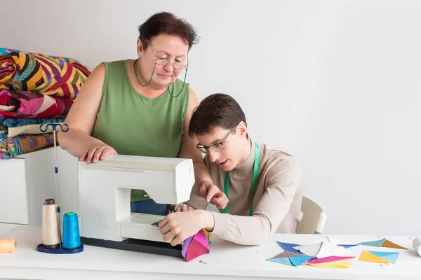 Colcha e retalhos de alfaiate masculino - uma mulher idosa no estúdio explica ao jovem como usar uma máquina de costura para a produção de tecidos de retalhos multicoloridos . — Fotografia de Stock