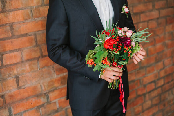elegant man dressed in a modern black suit and white shirt holding in hand a wedding flowers bouquet from red carnation. Close up color vertical image of male hands holding bunch of bright flowers.