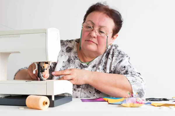 Retalhos e colchas na oficina de uma mulher alfaiate no fundo branco mulher mais velha com óculos está se preparando para trabalhar na máquina de costura com pedaços de tecidos coloridos . — Fotografia de Stock