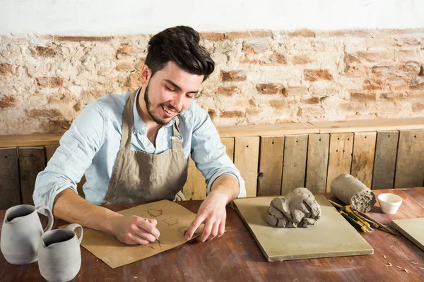 potter, workshop, artisan tools, ceramics art concept - smiling young male man working on the design of future products, a ceramist sitting behind desk with tools, fireclay and finished jugs.