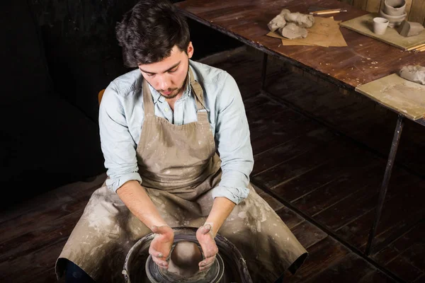 Potter, atölye, Seramik Sanat kavramı - genç adam el ile ham şamot potters tekerlek üzerinde bir erkek bir kap araçlarıyla, önlük ve gömlek, üstten görünüm ana ahşap masa yakınındaki sculpts.