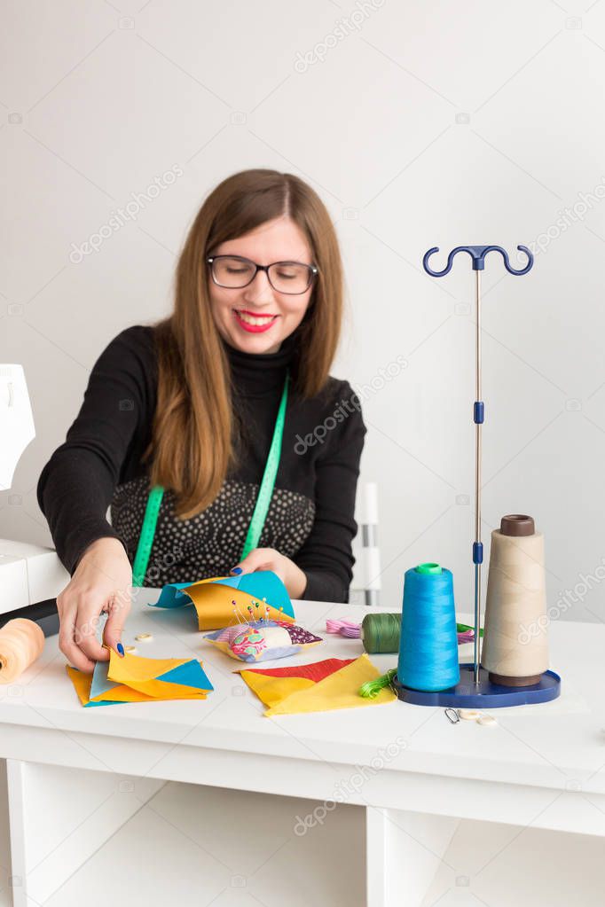 needlework and quilting in the workshop of a young woman, a tailor - smiling woman in glasses tailor at work with pieces of colored cloth on the table with threads, fabrics, needles