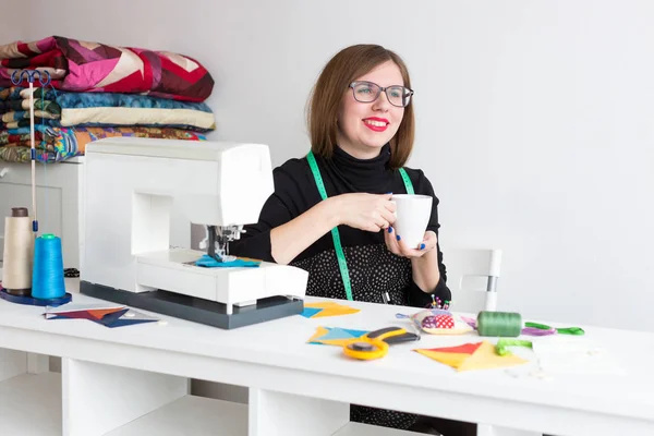 La costura y el acolchado en el taller del sastre - la interrupción del trabajo con la taza del té de la sastre hermosa joven a la mesa con la máquina de coser, las telas y los hilos — Foto de Stock