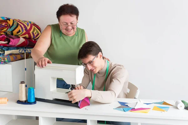 Colcha e retalhos de alfaiate masculino - uma mulher idosa no estúdio com um sorriso olhando para o trabalho dos jovens na máquina de costura para a produção de retalhos multicoloridos — Fotografia de Stock