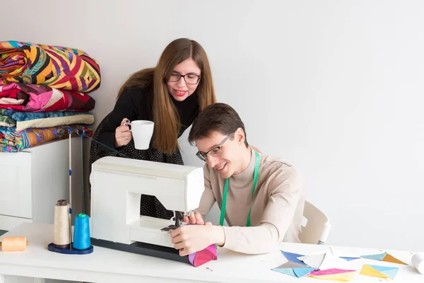 Acolchado y patchwork de sastre masculino - joven hermosa mujer en el estudio con la taza en la mano mirando el trabajo de un joven sastre hombres en la máquina de coser para la producción de telas patchwork multicolores . — Foto de Stock