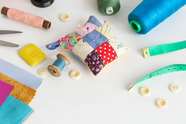 Needlework, craft, sewing and tailoring concept - tools closeup on white desk, measuring meter, thread spools, pieces of colorful patchwork fabric, pincushion, buttons, soap, scissors. — Stock Photo, Image