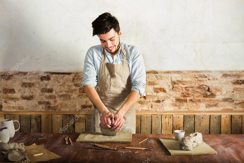 potter, workshop, ceramics art concept - standing young brunette man dressed in an apron, male hands knead the fireclay, a ceramist with raw materials on wooden table with sculpting tools set.