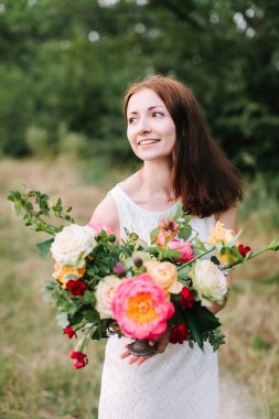 Güzellik, düğün, Bahar, doğa, dekorasyon konsepti - beyaz parti elbise renkli kokulu güzel peonies, gül ve karanfil oluşan koyu kahverengi saçlı düzgün gülümseyen kadın