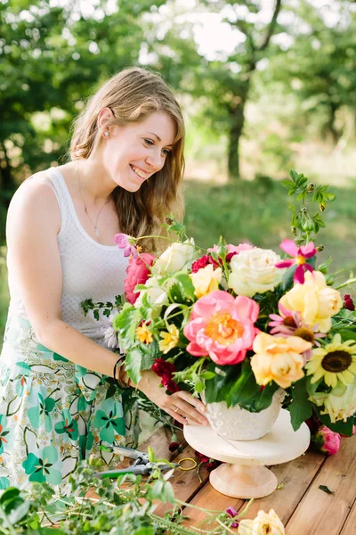 Хобби, работа, цветочный дизайн, свадьба, праздничная концепция - красивая улыбающаяся женщина в летнем платье с зеленым шрифтом и с легким макияжем, расставляющая розы, пионы и диафеты в большом букете — стоковое фото