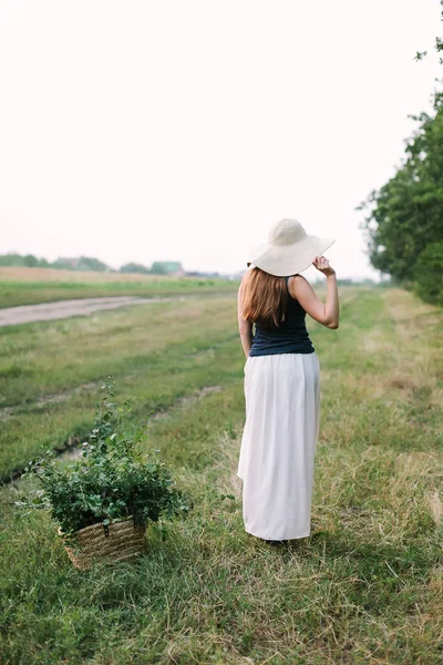 lifestyle, country, healthlife, nature concept - beautiful figure of woman in white maxi skirt and straw hat standing in field of grass, her handbag with oak branches lying nearby