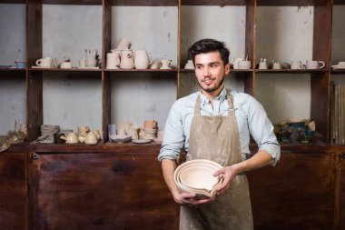 Potter, araçlar, Seramik Sanat kavramı - bir önlük tribünde ustası pişmiş cups ile işyerinde ve testiler, arka plan gülümseyen bitmiş mutfak eşyaları ile giyinmiş sakallı yakışıklı genç esmer erkek.