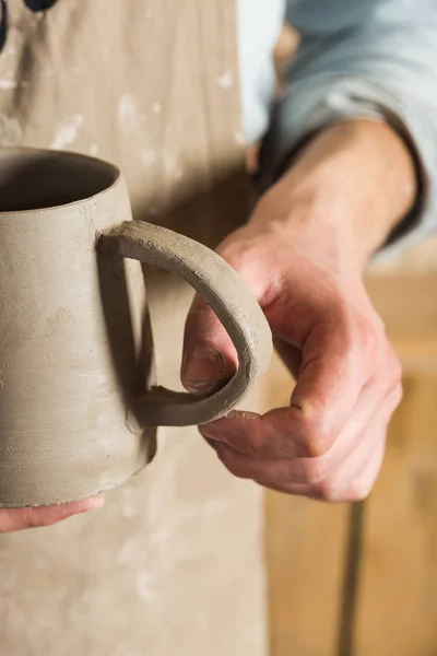 Cerâmica, oficina, conceito de arte cerâmica ceramista segurando copo de barro inacabado e examina cuidadosamente o produto, mestre masculino vestido com um avental de barro manchado, mãos artesanais segurando jarro não cozido . — Fotografia de Stock