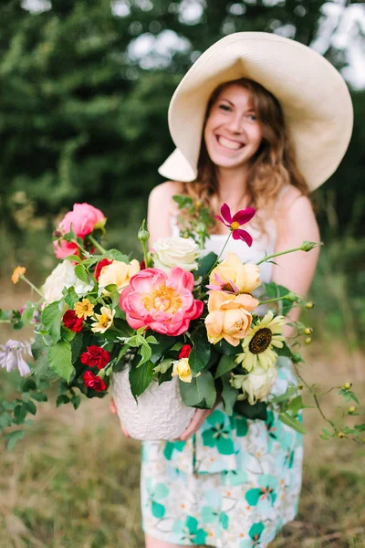 結婚式、花柄のデザイン、美しさ、カントリー ライフ コンセプト - 麦わら帽子とグランドの美しい牡丹、柔らかい黄色の雪崩、明るい赤いカーネーションの花束と夏のドレスで笑う女性を魅力的な — ストック写真