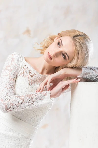 Vestido de novia de moda, hermoso modelo rubio, peinado de novia y concepto de maquillaje - joven mujer bonita en vestido blanco sentado en el interior sobre fondo claro, linda hembra posando en el estudio . — Foto de Stock