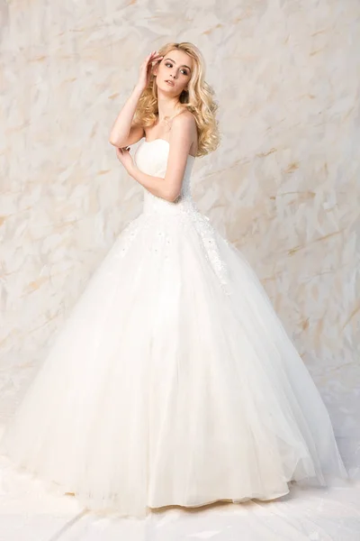 Vestido de moda, hermoso modelo rubio, peinado de novia y concepto de maquillaje - joven chica encantadora en vestido de fiesta de boda blanco, de pie en el interior sobre fondo claro, mujer esbelta romántica posando . — Foto de Stock