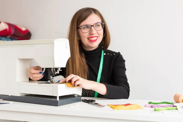 Handarbete och quiltning i verkstaden för en ung kvinna, en skräddare på vit bakgrund - en ung flicka med ett leende på hennes ansikte sy på symaskin — Stockfoto