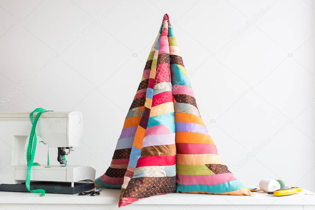 quilting - high pillow in the form of a pyramid of colorful quilted fabrics made of pieces of patchwork fabrics on the table next to the scissors, scraps of fabrics, coils of thread, sewing machine.