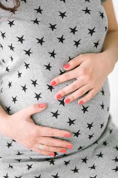 Těhotenství, ženskost, narození dětí koncepce - detailní na kulaté břicho mladé budoucí matky, ruce s růžová manikúra nastávající matky jemně podporují žaludku, šedé šaty s vzor-hvězdy. — Stock fotografie