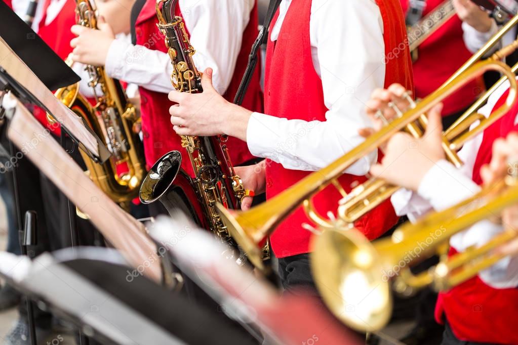 jazz band music performance concept - orchestra of wind instruments during the concert, selective focus on hands of musicians playing on trumpets and saxophones, closeup male in red costumes.