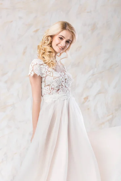 Vestido de novia de moda, hermoso modelo rubio, peinado de novia y el concepto de maquillaje impresionante mujer joven en vestido blanco de lujo en el interior sobre fondo claro, posando bastante femenina con una sonrisa . — Foto de Stock