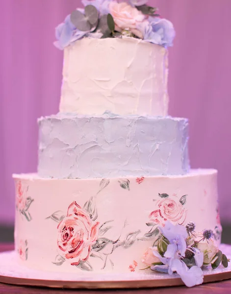 bakery, celebration, engagement concept. wonderful three-tiered wedding cake in light tender pastel shades decorated with painted roses on the first tier and natural flowers on the top