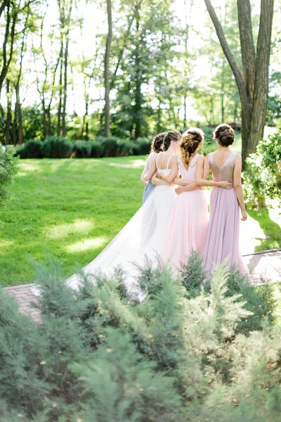 stock image celebration, engagement, woman frienship concept. backs of four young slim women, happy bride in traditionaly white dress and her bridemaids, they are walking on the park full of trees and bushes