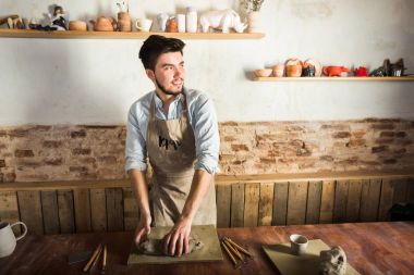 Potter, atölye, Seramik Sanat kavramı - duran genç gülümseyen esmer adam elleriyle yoğurmak şamot, önlük, bir Fransız araçları kümesi heykel ile ahşap masa üzerinde hammadde ile giymiş erkek.