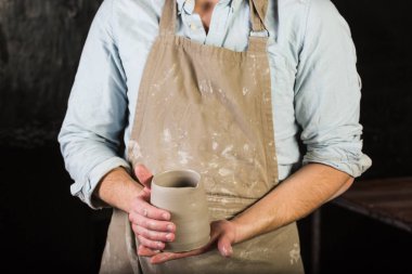 çanak çömlek, taş, Seramik Sanat kavramı - unfired kil, genç adam kirli önlüğü standında bir atölye fincan holding üzerinde ana eller erkek taze bir ürün daha fazla işleme önce inceliyor yakın çekim.