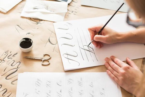 Diseño gráfico, escritura a mano, concepto de creación. tiernas manitas de pintora que inscriben letras ornamentales decoradas en el papel forrado blanco con la ayuda de la punta delgada del pincel y la tinta negra —  Fotos de Stock