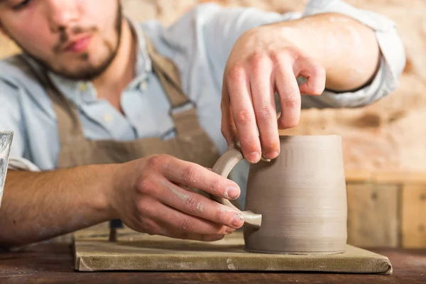 Oleiro, grés, conceito de arte de cerâmica - mãos artesanais conectam o copo e o punho feito de argila crua, dedos mestres trabalham com pedaços de argila, o macho senta-se em uma oficina atrás da mesa . — Fotografia de Stock