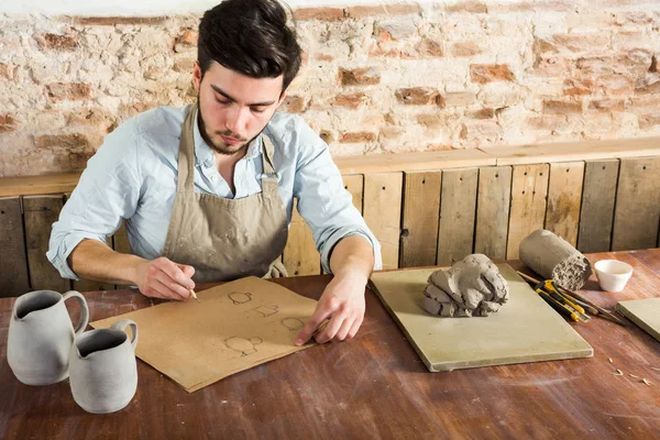 potter workshop, artisan tools, ceramics art concept - young man works with design of future utensil products, a ceramist sitting behind desk near tools, fireclay and finished pitchers.