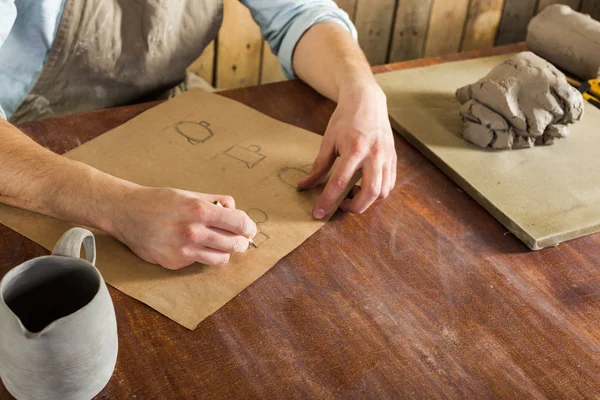 Fabrication de poterie, outils artisanaux, concept d'art céramique vue du dessus des mains des jeunes hommes au travail sur le dessin de la faïence, un céramiste vêtu d'une chemise et d'un tablier assis derrière le bureau et une cruche finie . — Photo