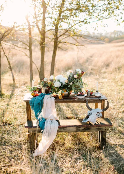 table setting, fall, travelling concept. on the wooden table for picnic standing in the field there is stunning bouquet of white flowers and cultery, decorative skull with horns placed on bench