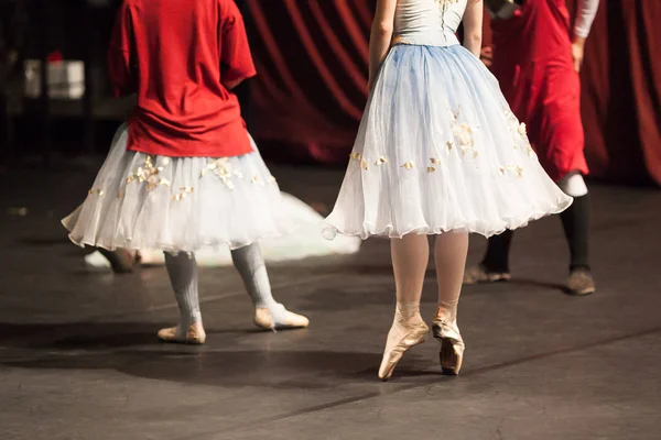 Dança, ensaio, conceito de bastidores. grupo de bailarinos que se alongam antes do concerto e uma das bailarinas ainda vestindo aquecedores de pernas cinza e grande camisola vermelha brilhante — Fotografia de Stock