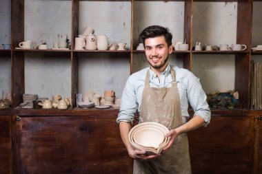 Potter, araçlar, Seramik Sanat kavramı - bir önlük ile bitmiş mutfak eşyaları ayakta, cups ile işyerinde ustası ve testiler, arka plan gülümseyen ana yakışıklı genç esmer erkek giymiş mutlu.