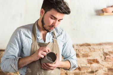 potter, atölye, Seramik Sanat kavramı - kil lekeli bir önlük giymiş ve bir işyerinde, genç craftsmans eller duran erkek Fransız çiğ kil sürahi ve araçları tutuyor.