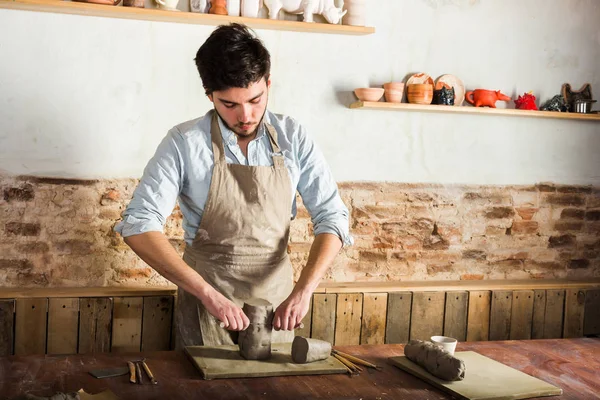 potter, kil atölye, Seramik Sanat kavramı - ayakta esmer adam erkek elleri dize kesme kil ile Fransız araçları heykel ile ahşap masa üzerinde hammadde ile bir önlük giymiş ayarla.