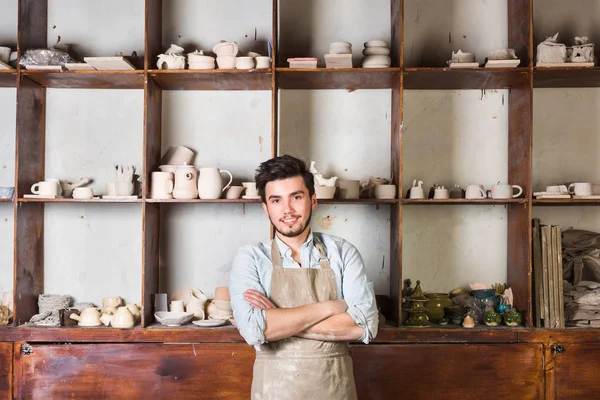 potter, tools, ceramics art concept - a handsome young brunette with a beard dressed in an apron, smiling male master ceramist at workplace with finished jugs and cups background.