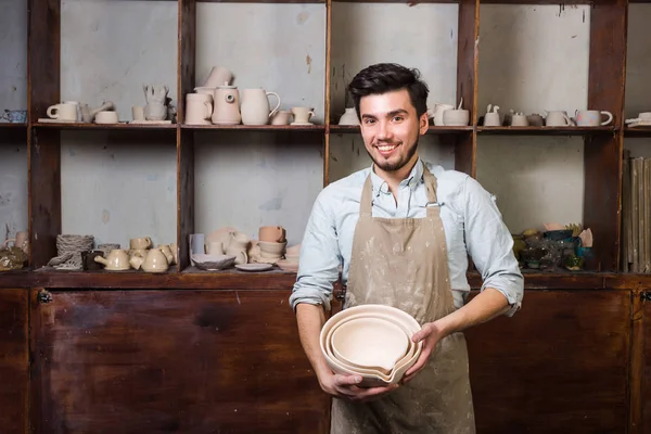 Potter, araçlar, Seramik Sanat kavramı - bir önlük ile bitmiş mutfak eşyaları ayakta, cups ile işyerinde ustası ve testiler, arka plan gülümseyen ana yakışıklı genç esmer erkek giymiş mutlu.