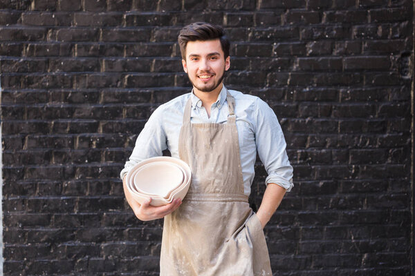 pottery, utensils, ceramics art concept - smiling male ceramist with hands holding some bowls, cute finished clay products and happy craftsman on the background of dark gray brick wall.