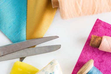 Dikiş, yamalama, terzilik ve moda konsepti - atölyedeki beyaz çalışma masasında yakın çekim araçları, makas, mavi ve pembe iplikler, iğneleme, renkli yama kumaş parçaları, dikey.