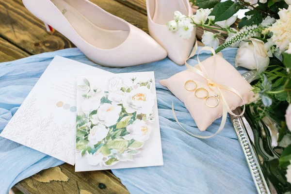 Style, wedding, tenderness concept. on the wooden table covered with light blue draping there is tidy pillow with golden rings, elegant pointed shoes and beautiful postcards — Stock Photo, Image