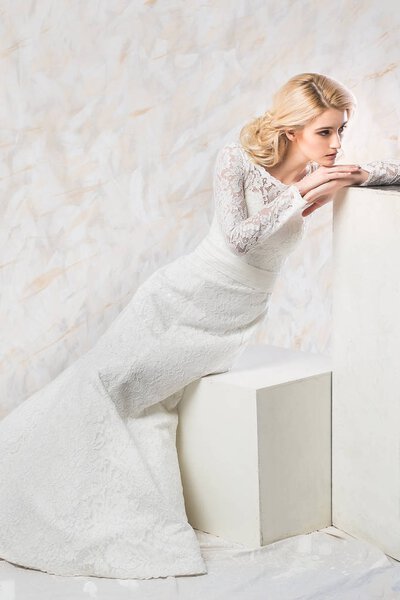 fashionable wedding dress, beautiful blonde model, bride hairstyle and makeup concept - young woman in luxury white gown sitting indoors on light background, pretty female posing in the studio.