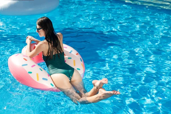 Servet, tatil, yaz konsepti. Koyu yeşil mayo ve güneş gözlüğü takmış, uzun siyah saçlı gülümseyen kadın havuzda şeffaf suyla birlikte kauçuk halkada yüzüyor.