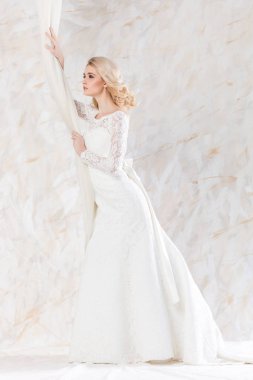 şık beyaz bir elbise, güzel bir sarışın manken, gelin saç stili ve makyaj konsepti - açık arka planda duran şirin gelinlikli zayıf genç kadın, perdelerin yanında poz veren çekici kız