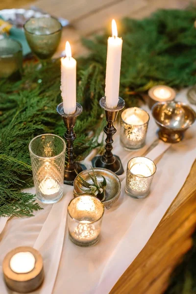 Weekend, date, silverware concept. holiday decor of table consists of amount of candles in varied holders of glass and metal, in the center of table there are lots of green conifers branches — Stock Photo, Image