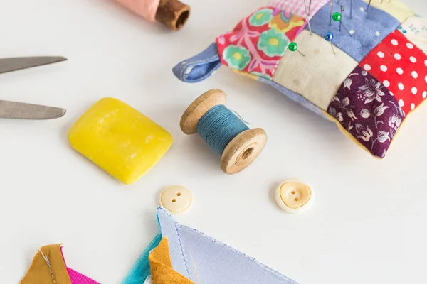 Sewing, patchwork, tailoring and fashion concept - closeup tools on white desk in studio, pincushion, blue and pink threads spools, white buttons, scissors, soap, pieces of colored patchwork fabric. — Stock Photo, Image