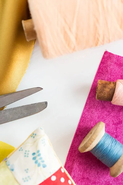 Dikiş, yamalama, terzilik ve moda konsepti - atölyedeki beyaz çalışma masasında yakın çekim araçları, makas, mavi ve pembe iplikler, iğneleme, renkli yama kumaş parçaları, dikey.