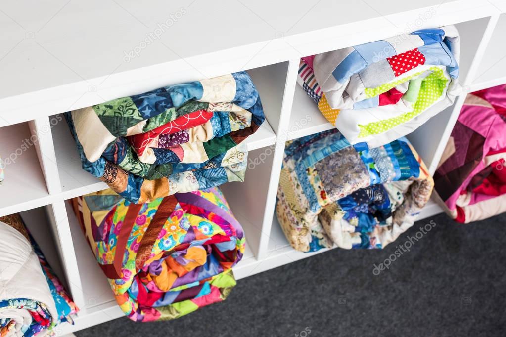 patchwork, sewing and fashion concept - colorful finished quilts in the studio at white shelves with few storage compartments, the warehouse of finished products, top view.